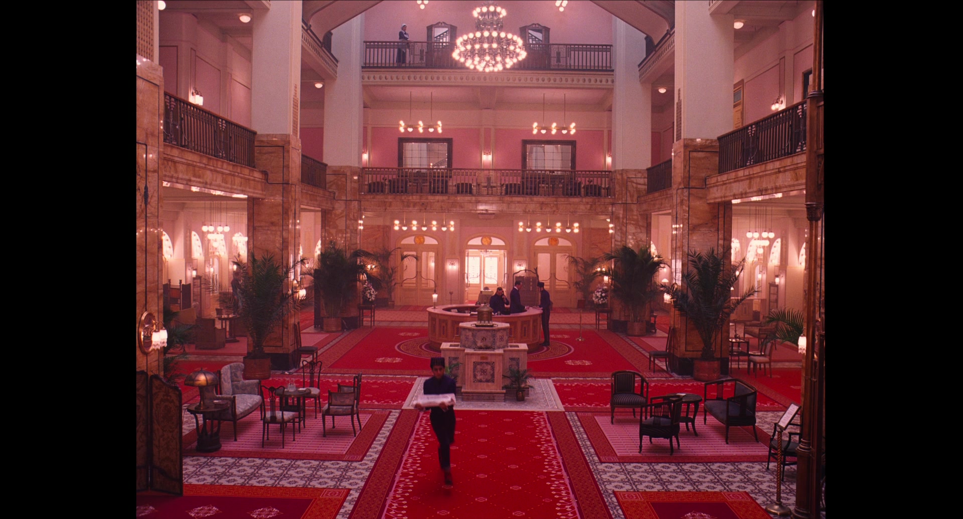 What Is Art Nouveau? Definition, Style, Artists & Film Influence 7 A wide, symmetrical view of the Grand Budapest lobby with red carpet runners, a central fountain, a circular reception desk, balconies with dark railings, pink walls, chandeliers, and a uniformed porter crossing the foreground.