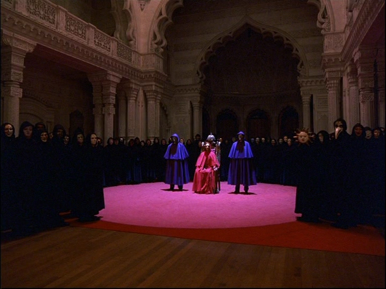 What is Mise-en-Scéne? Definition, Elements & Examples 3 A masked figure in red sits on a throne surrounded by robed individuals in a grand, dimly lit hall with detailed arches and a red carpet.
