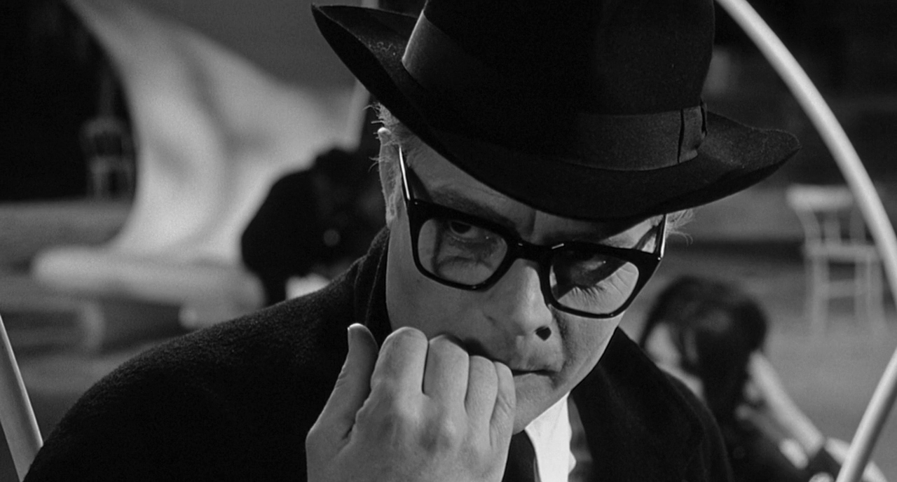 30 Films Every Budding Film Student Should Watch & Why 7 A man in a hat and glasses bites his finger while staring intensely, with blurred background figures behind him.