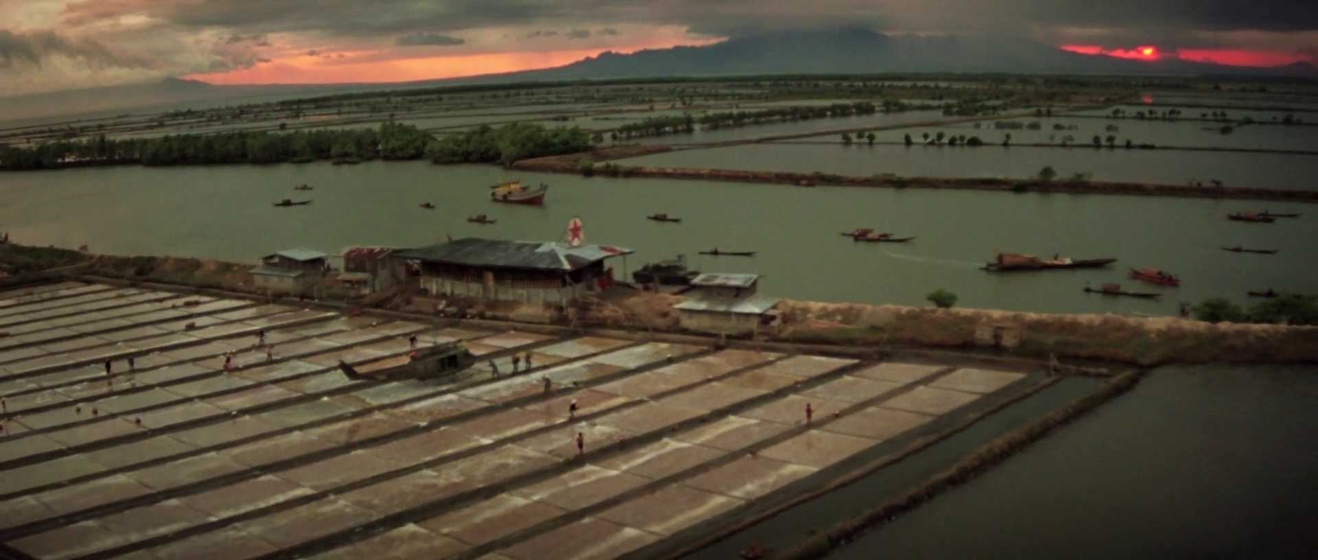 Context in Screenwriting and Film: A Complete Guide 3 A wide aerial view of rice paddies, rivers, and small boats at sunset, with a helicopter landed near soldiers and buildings