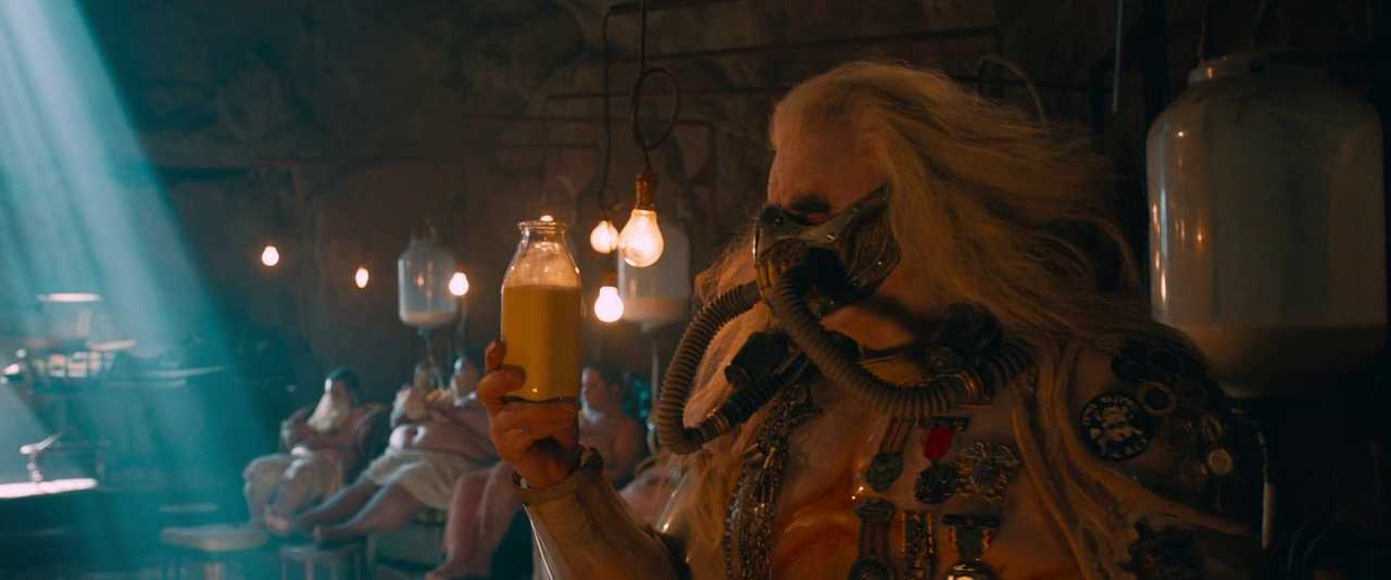 What is Exposition in Film? Definition, Types, and Techniques 3 A masked, long-haired War Boy holds up a glass bottle of milk in a dim room, with several women lying in the background connected to pumping equipment.