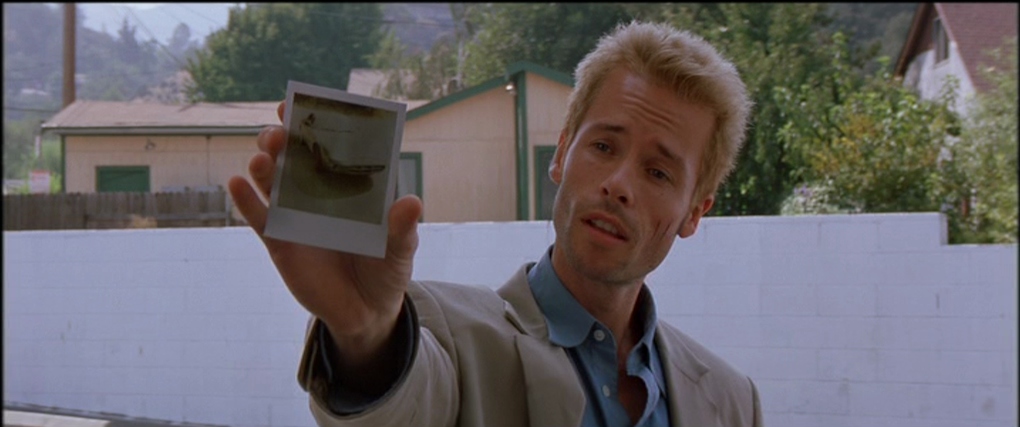 Context in Screenwriting and Film: A Complete Guide 4 A man with bleached hair holds a Polaroid photo in front of him while standing outside near white buildings and trees