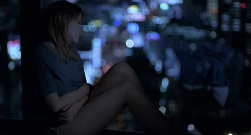 What Is the Setting in Film? Time, Place & Mood 2 A woman sits with knees pulled to her chest at a hotel window while out-of-focus city lights glow outside at night.