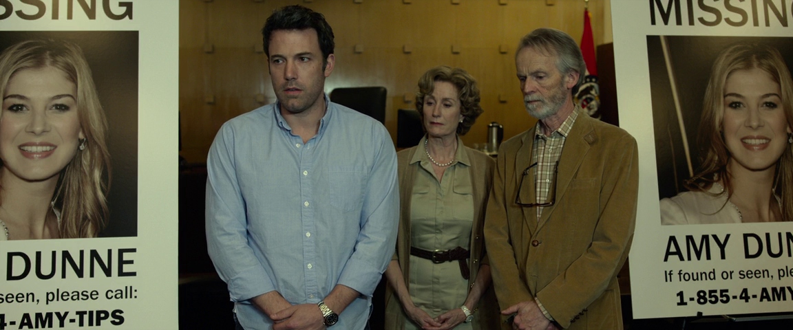 What is a Red Herring? Meaning & Examples from Film & Literature 4 Nick Dunne stands in the center of a room between two large “Missing” posters showing Amy’s face, with an older woman and man behind him.