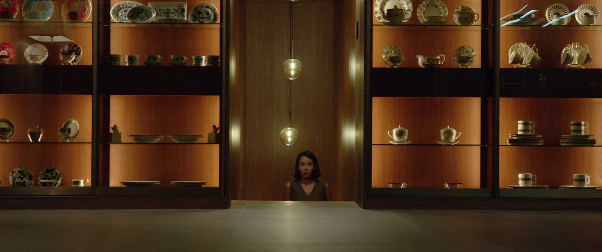 Theme in Film: Definition, Meaning, and Scene Examples 5 A wide, centered shot of a woman standing low in a doorway-like opening between two lit display cabinets filled with plates and teacups, with two hanging lights above her.