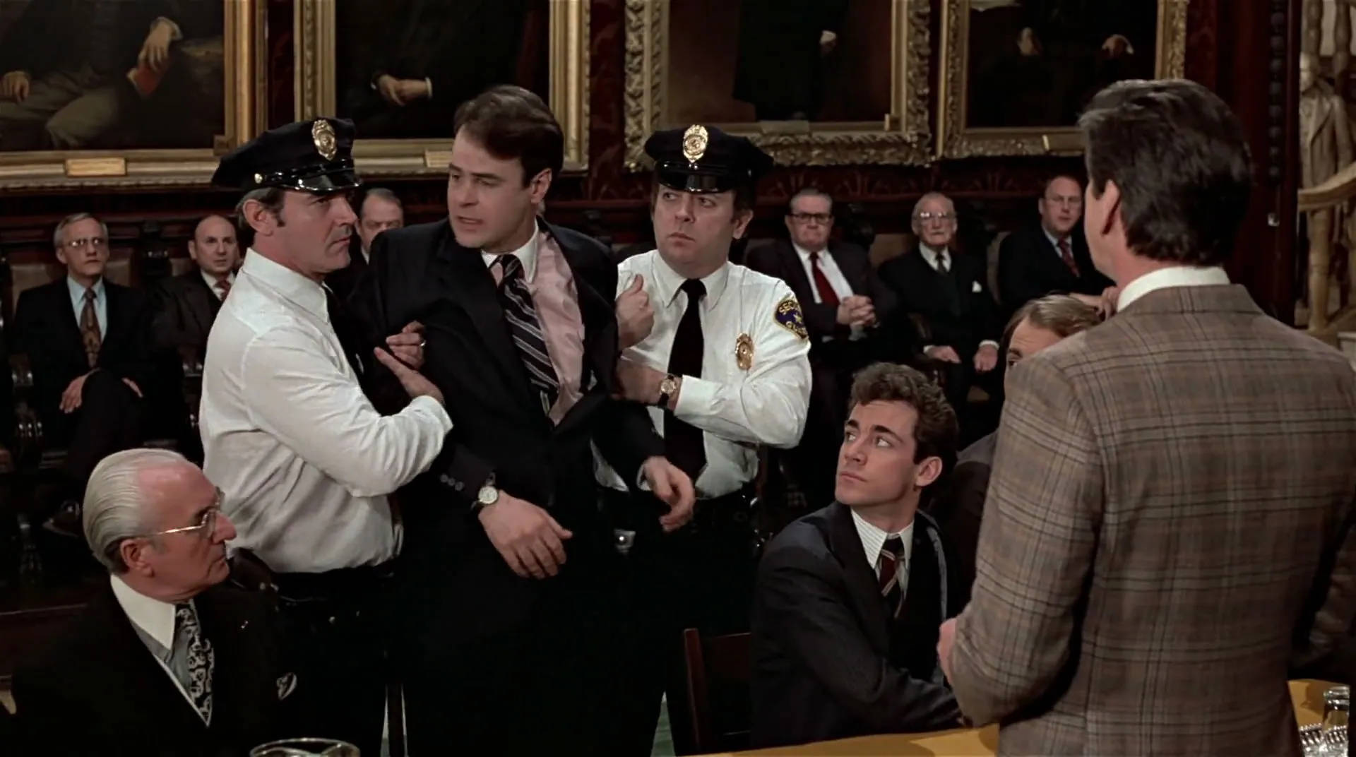Peripeteia in Film. Definition, Meaning & Examples 6 Two uniformed officers restrain Louis Winthorpe III in a crowded wood-paneled room filled with men in suits.