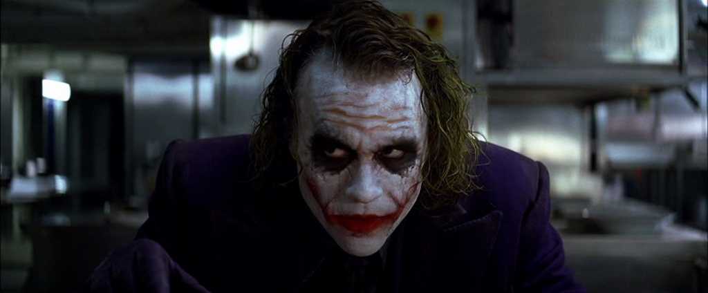 What Is a Stock Character? Definition & Film Examples 3 Close-up of the Joker leaning forward over a table in a stainless-steel room, staring intensely.