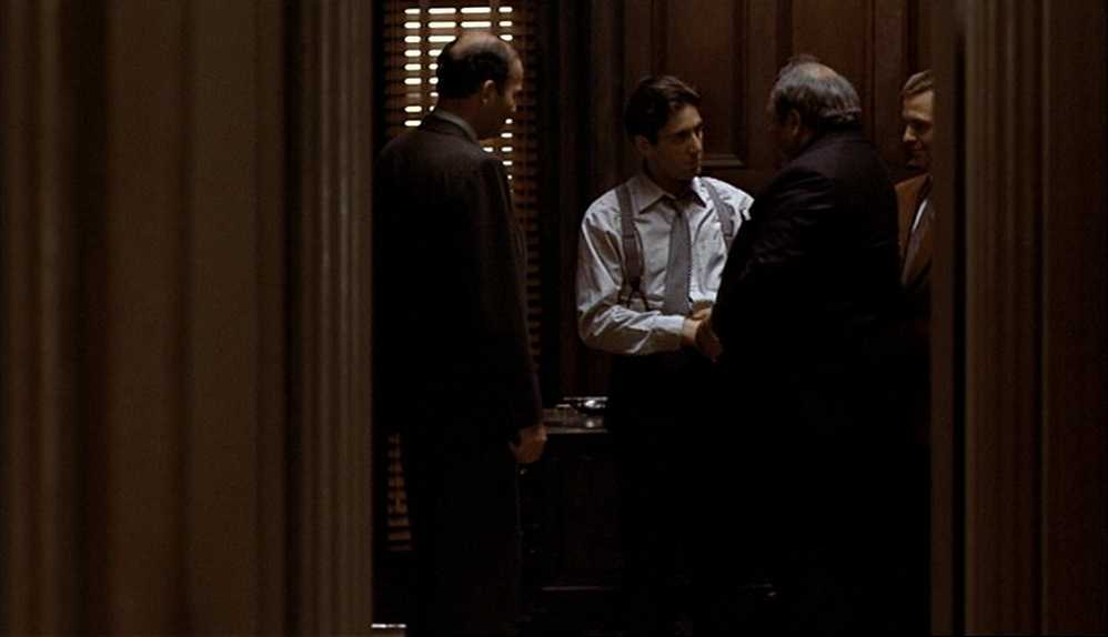 What Is a Character Arc? Definition, Stages & Film Examples 3 Michael Corleone stands in a narrow, dim hallway with three suited men around him, lit by slatted light from venetian blinds.
