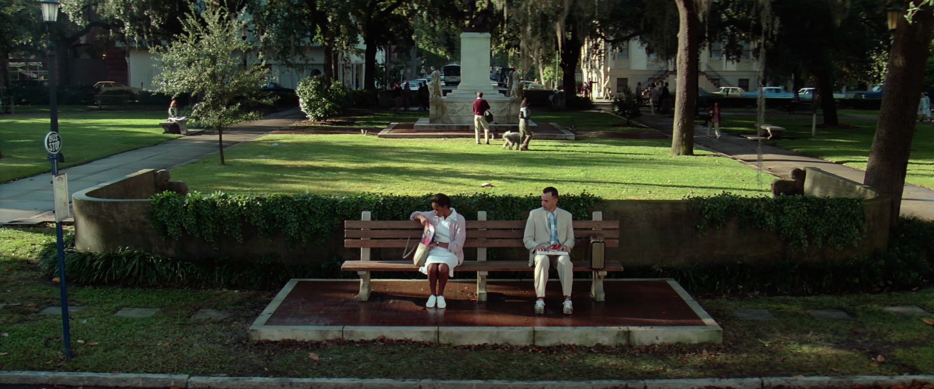 Analogy Explained: Meaning, Function + Film Examples 4 A man in a light suit sits on a park bench beside a woman at a bus stop, holding a small box on his lap in a sunlit green square.