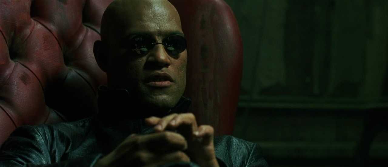 Analogy Explained: Meaning, Function + Film Examples 5 Morpheus sits back in a red leather chair in a dim room, wearing dark sunglasses and black clothing, with his hands clasped as he speaks.