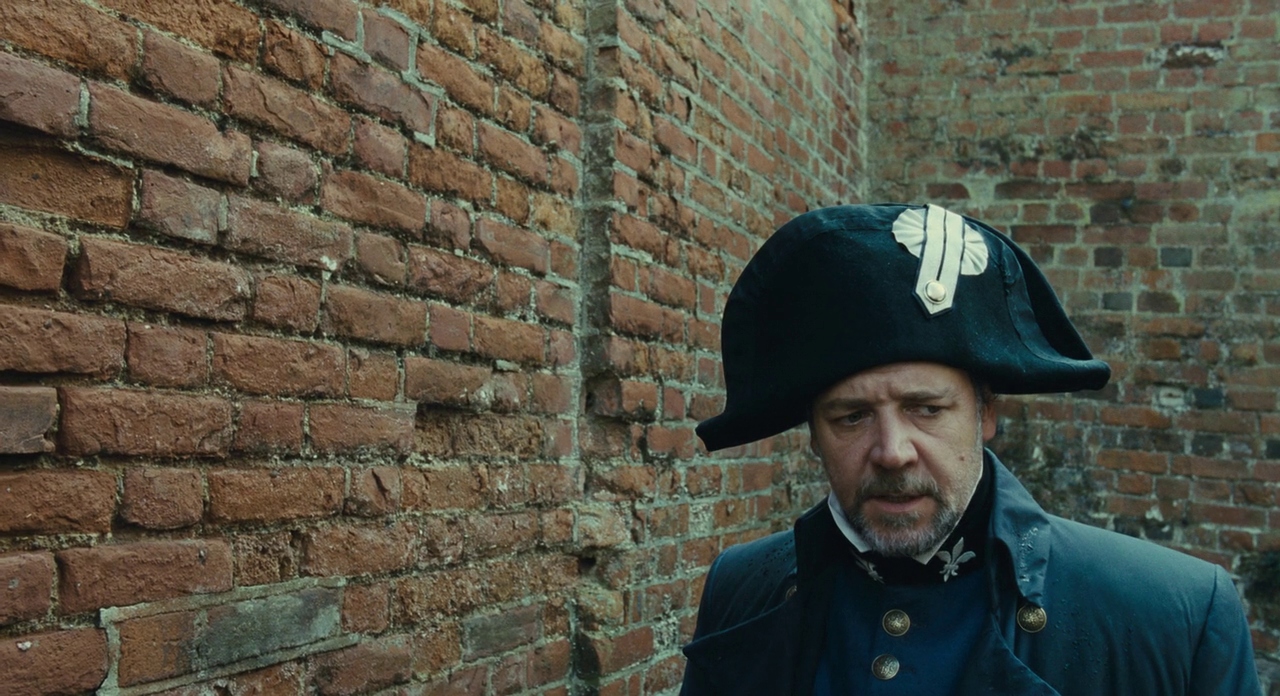 What is an Anti-Villain? Definition & Examples from Film 5 Inspector Javert in a dark uniform and bicorne hat walks beside a wet brick wall, looking down with a tense expression.