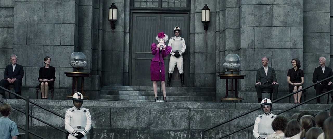 What is a Catalyst in Film? Definition & Examples 2 Wide shot of the Reaping stage with Effie Trinket in a bright pink outfit at a microphone, Peacekeepers in white uniforms, and seated officials on either side in front of a large stone building.