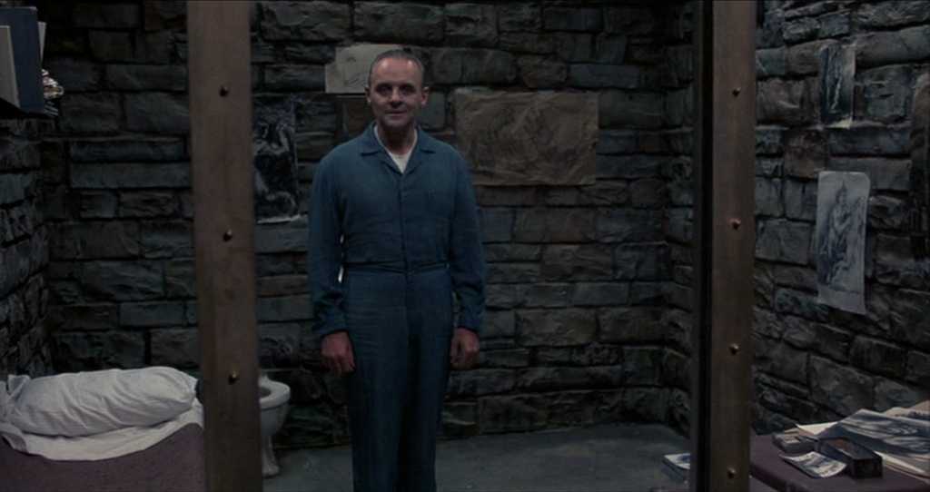 What is a Static Character? Definition & Film Examples 4 Hannibal Lecter stands centered in a dim stone prison cell wearing a blue jumpsuit, with a bed and toilet on the left and drawings and papers on the walls and table.