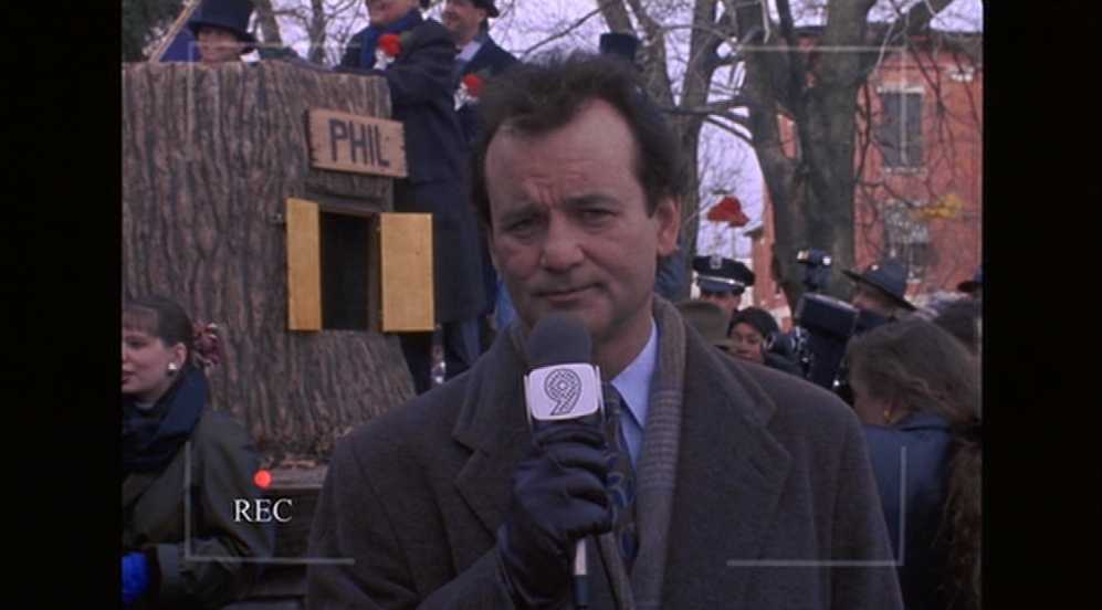 What is a Dynamic Character? Definition & How to Write One 3 Phil Connors holds a TV microphone at a Groundhog Day crowd event, with a stump labeled “PHIL” behind him and a camcorder “REC” overlay on the image.
