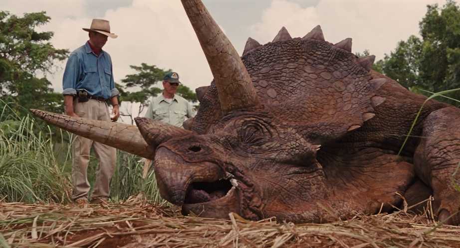 What is Verisimilitude in Film? Definition, Types & Examples 2 Two men in outdoor clothing stand in tall grass beside a large triceratops lying on the ground with its eyes closed.