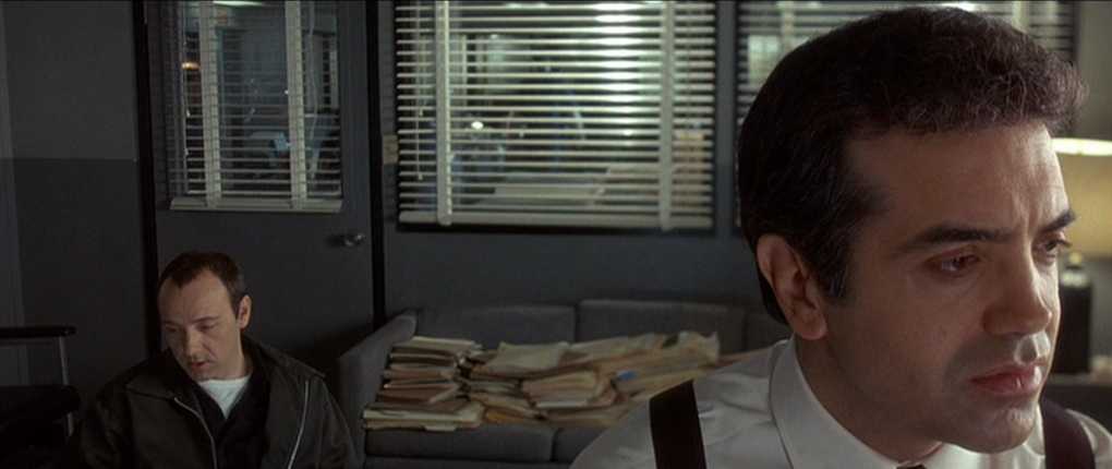 Narrator Types Explained. Definition and Examples from Film 6 Close-up of a serious man in a white shirt and suspenders in the foreground, with another man seated in the background inside a small office with blinds, a couch, and stacks of files.