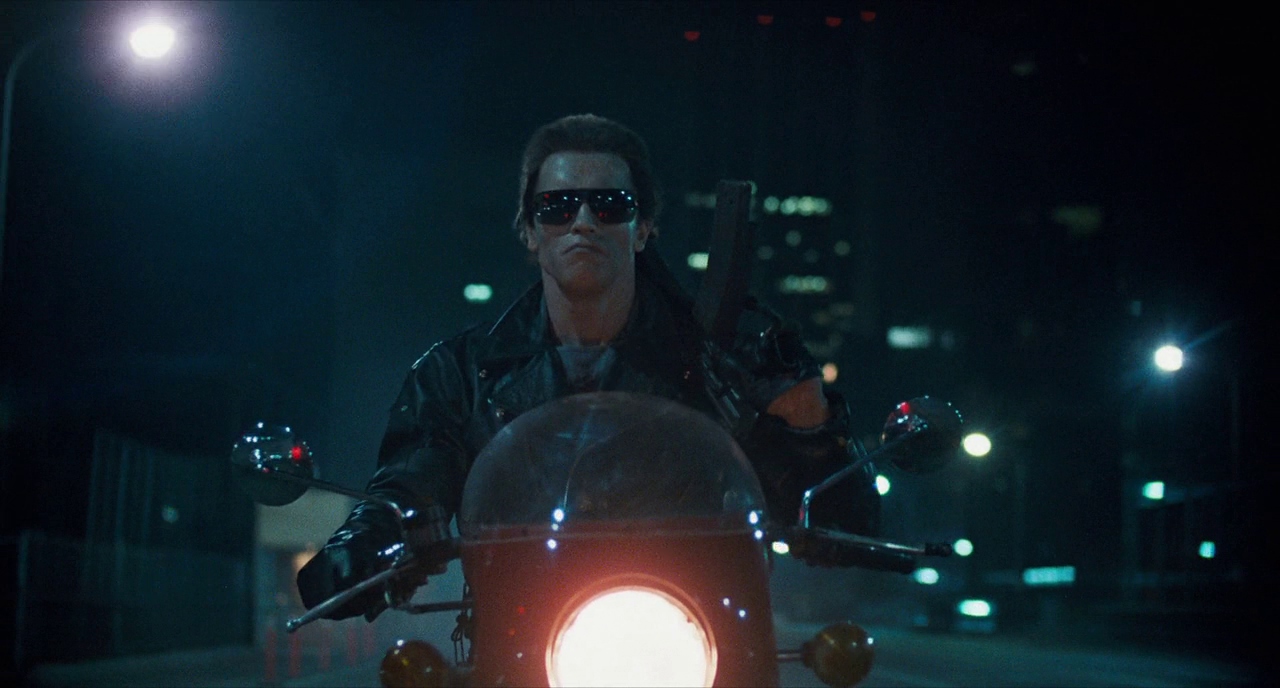 What is a Flat Character? Definition & Examples from Film 3 The Terminator rides a motorcycle at night with city lights behind him, wearing sunglasses and a black leather jacket with a weapon strapped over his shoulder.
