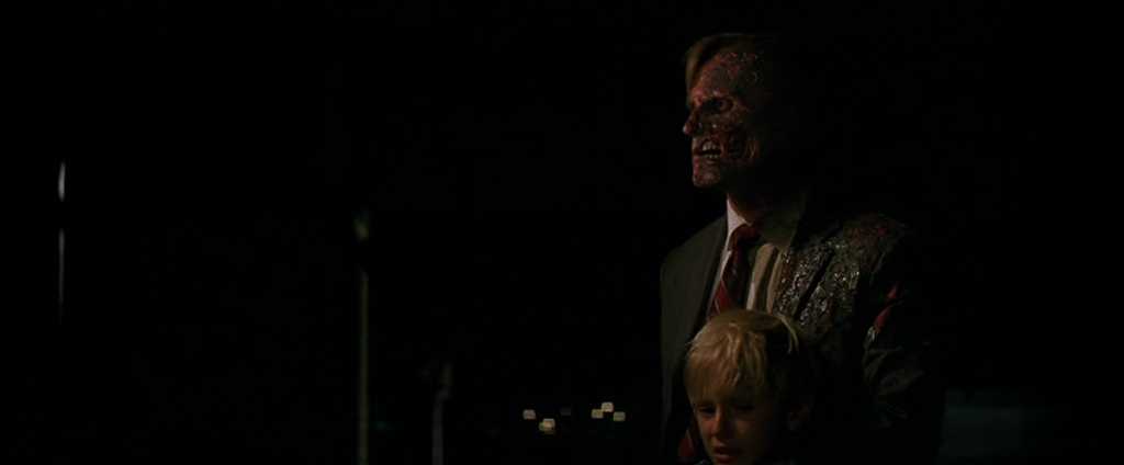 What is a Round Character? Definition & Film Examples 2 A suited man with half his face burned stands in the dark at night while holding a blond child close in front of him; distant city lights glow in the background.