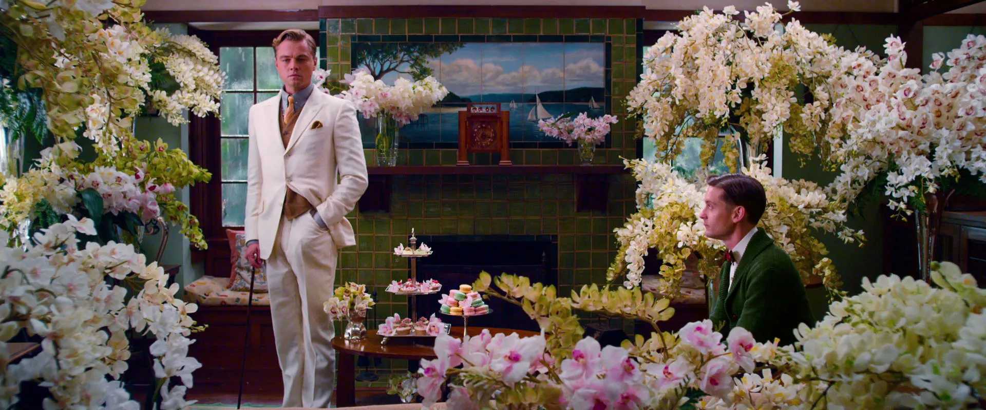 What is a Round Character? Definition & Film Examples 4 Gatsby in a white suit stands near a fireplace in a flower-filled room, while Nick sits to the right in a green sweater; white blossoms and pastel flowers crowd the frame around them.