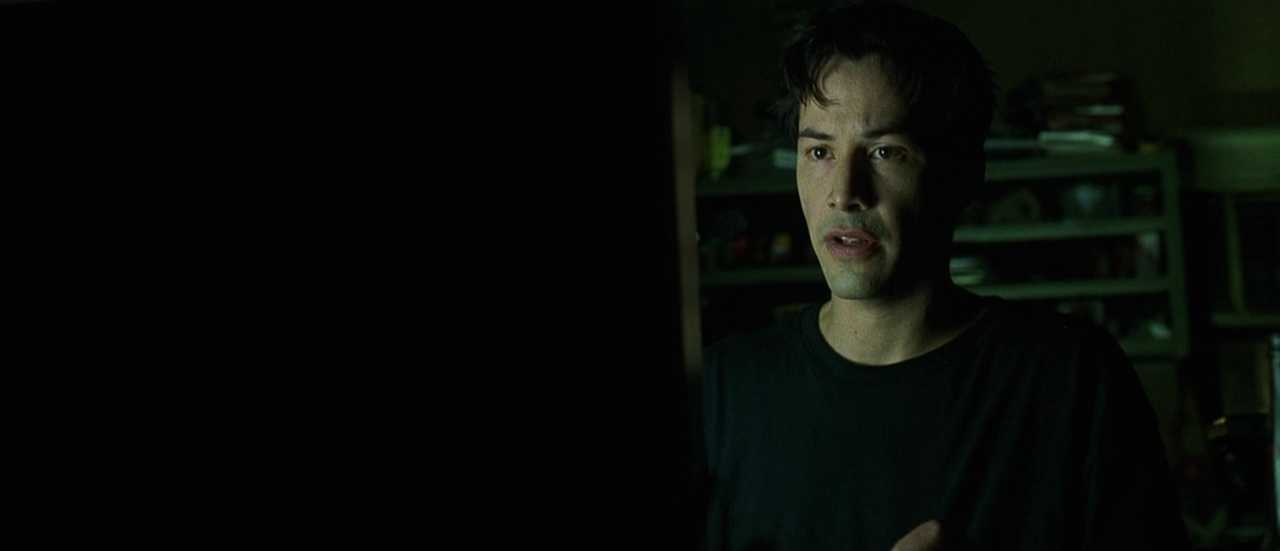 What is Verisimilitude in Film? Definition, Types & Examples 4 Neo sits in a dim, green-lit room with shelves behind him, looking tense as he listens and tries to understand what is happening.