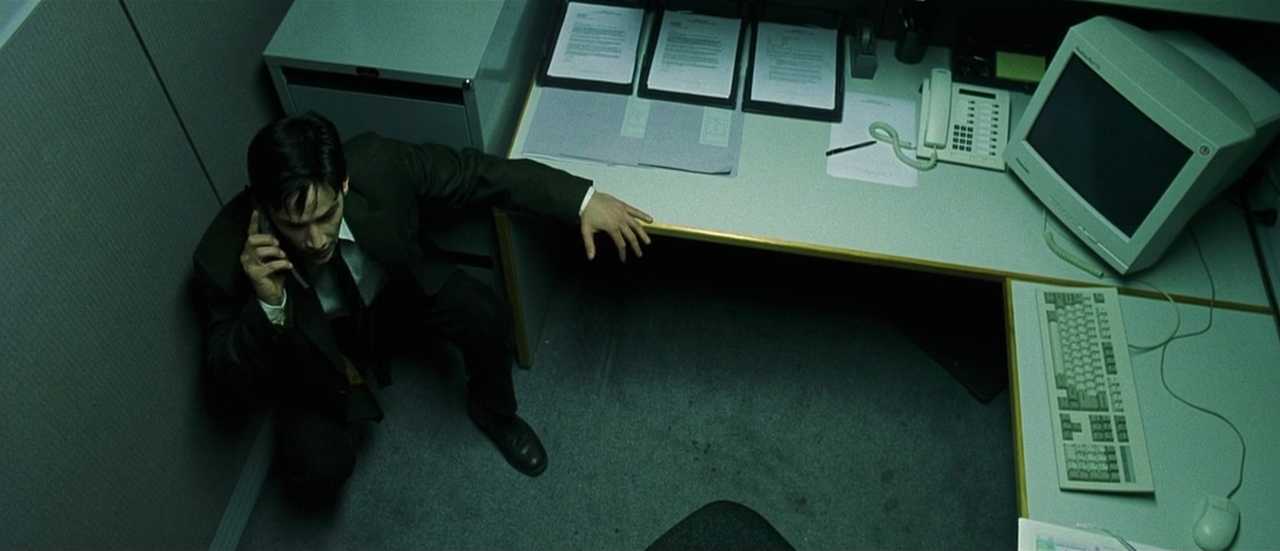 What is a Catalyst in Film? Definition & Examples 4 High-angle shot of Neo crouched in the corner of his office cubicle, holding a phone beside a desk with a CRT monitor, keyboard, and scattered papers.
