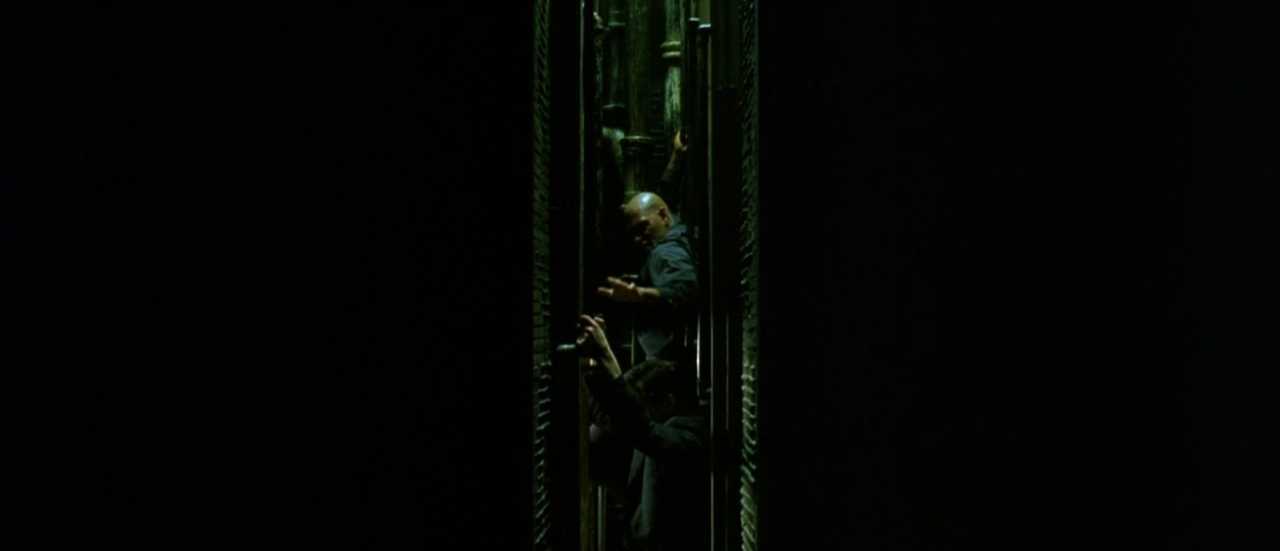 What is Verisimilitude in Film? Definition, Types & Examples 6 Two people climb through a narrow vertical gap inside a dark, green-tinted high-rise interior, pressed between walls and metal structures.