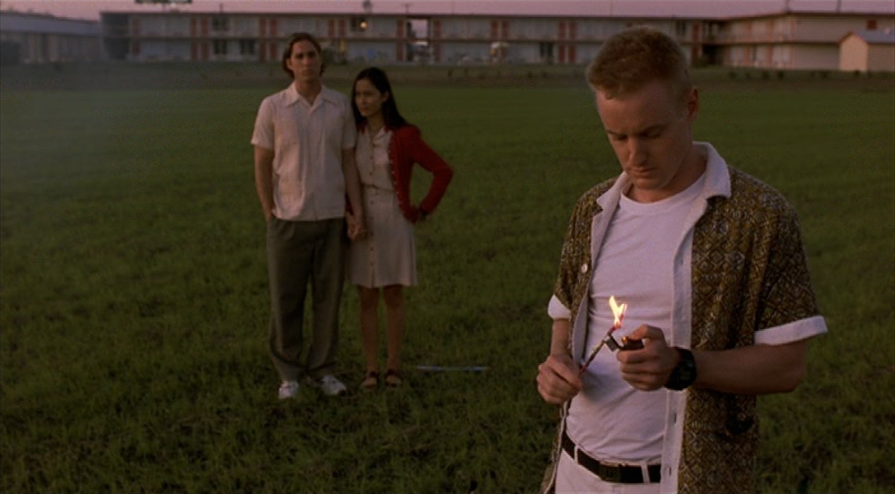 Guide: Best Wes Anderson Movies (For Filmmakers to Learn From) 8 A man lights a firework in the foreground while a couple stands in the distance on a grassy field in Bottle Rocket