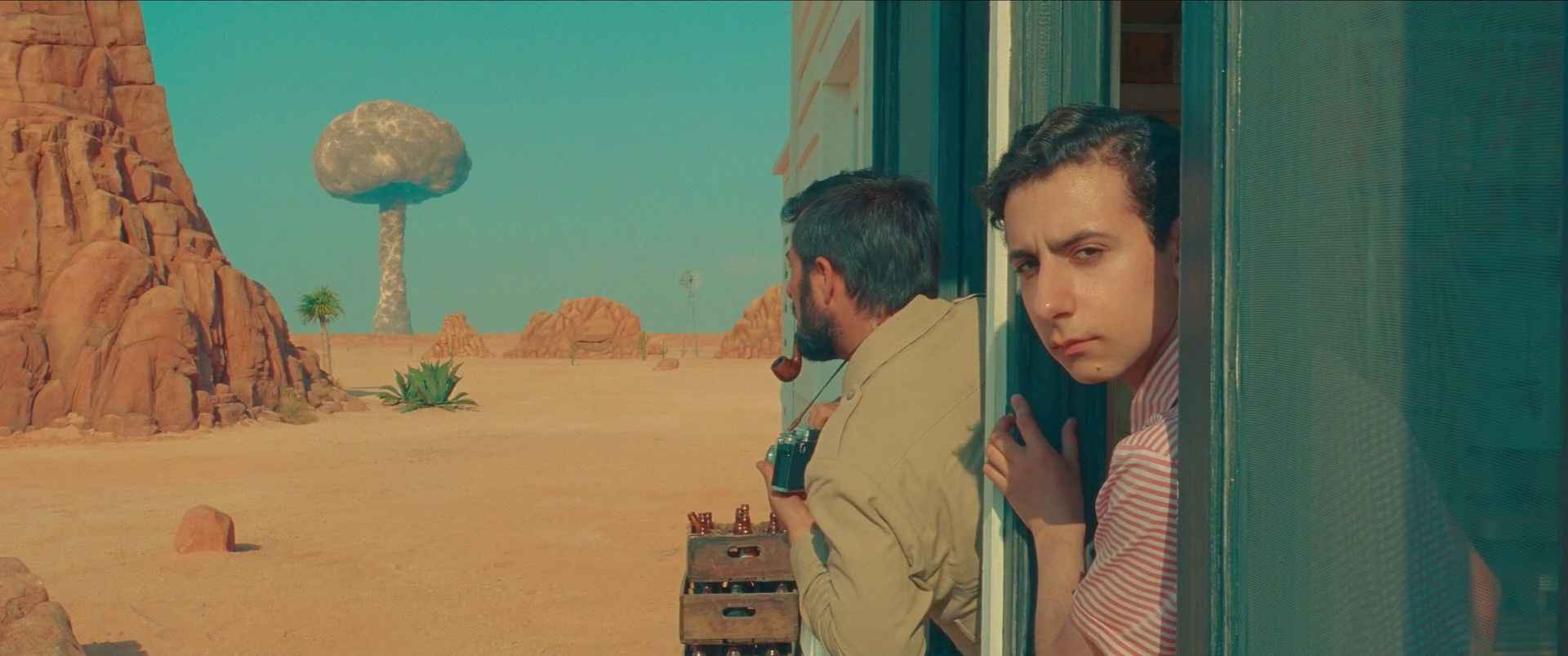 Guide: Best Wes Anderson Movies (For Filmmakers to Learn From) 2 Two characters peek out of a doorway in the desert as a mushroom cloud rises in the distance in Asteroid City
