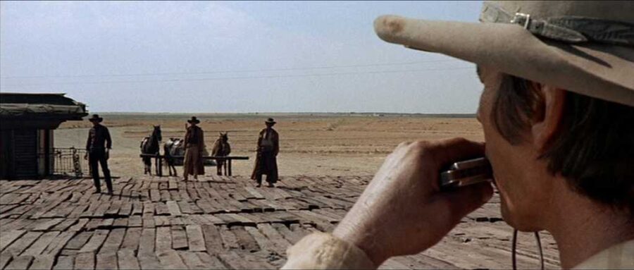 What Is Eurocult Cinema? Definition, Directors & Legacy 5 Man playing harmonica watches three gunmen across wooden platform