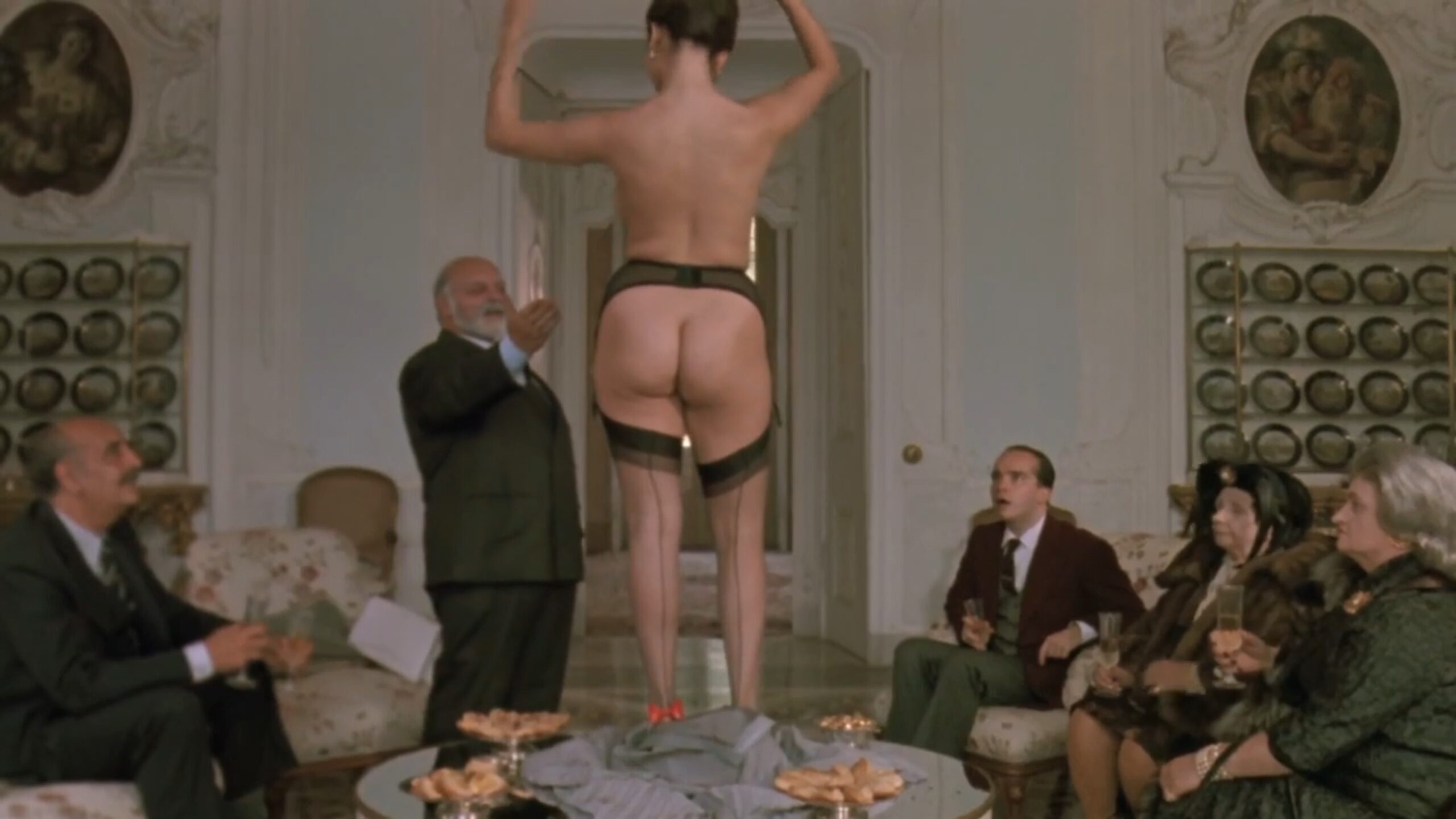 Tinto Brass Film Style: Sex, Humor, and Italian Genre 5 Woman in stockings standing on a table while a group of guests watches, showing silly behavior and exaggerated roles in a playful, risqué scene.