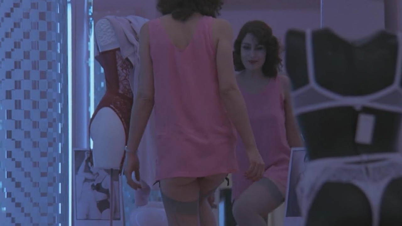 Sex, Power, and Rebellion: Symbolism in Tinto Brass Movies 9 Woman in pink dress looking at herself in a mirror inside a lingerie store, with mannequins and soft blue lighting suggesting introspection and sensuality.