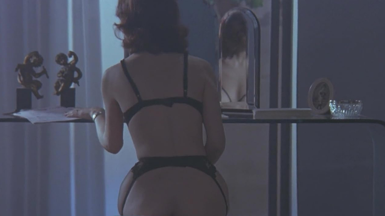 Sex, Power, and Rebellion: Symbolism in Tinto Brass Movies 4 Woman in black lingerie leaning over a table, seen from behind, with her reflection visible in a mirror, emphasizing voyeuristic framing in a dimly lit room.