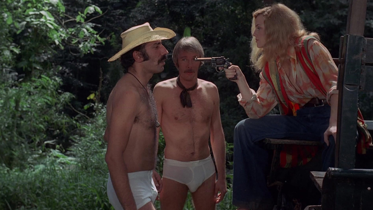 What Is Exploitation Cinema? Definition & Key Examples 13 Woman sitting on a jeep points a revolver at two shirtless men in underwear in a jungle setting in The Big Doll House (1971).