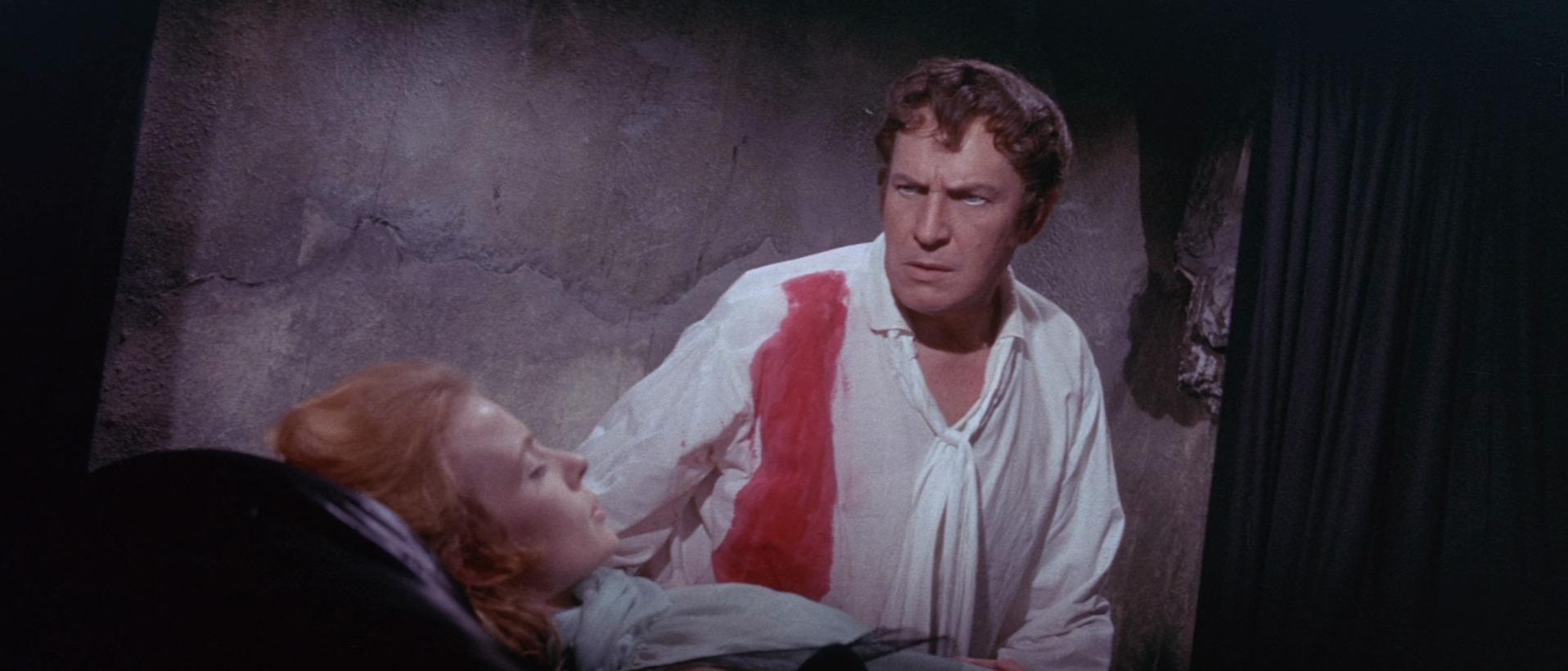 What Is Exploitation Cinema? Definition & Key Examples 12 Vincent Price in The Tomb of Ligeia (1964) wearing a torn white shirt with a red stain, sitting beside a sleeping woman in a stone-walled room.