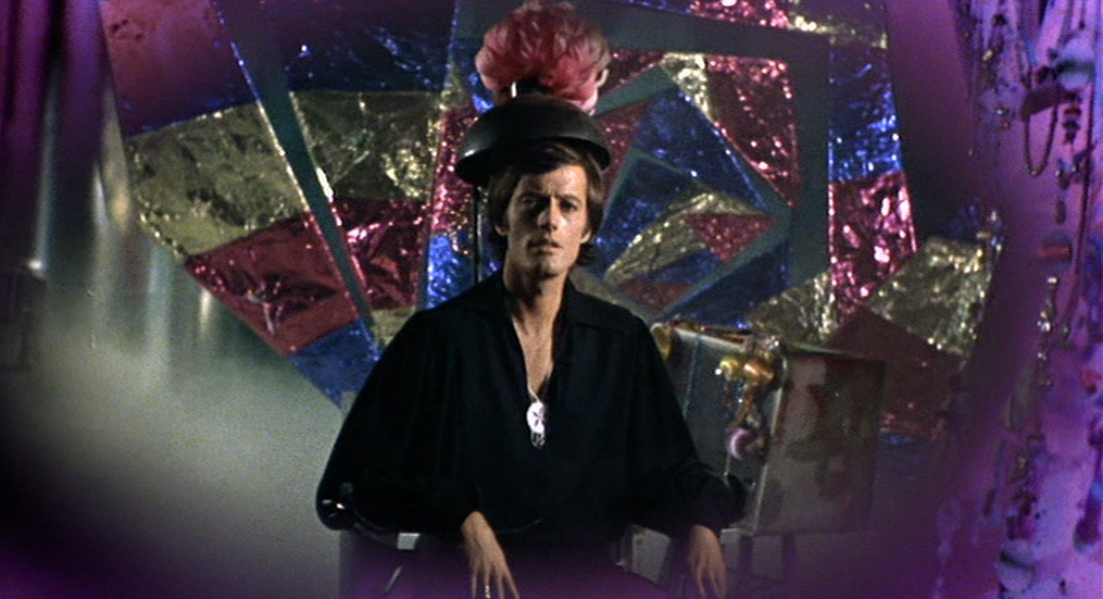 What Is Exploitation Cinema? Definition & Key Examples 11 Man sitting in front of colorful psychedelic shapes in The Trip (1967), wearing a feathered hat and black shirt.