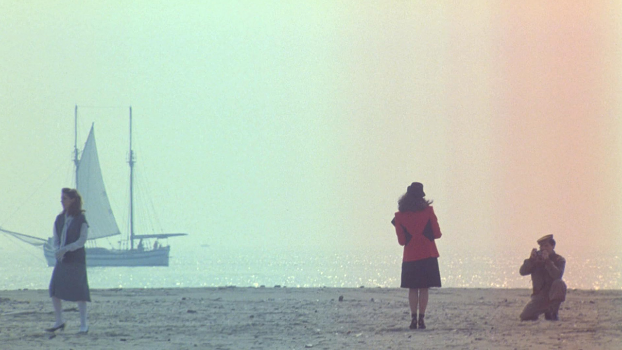 Tinto Brass Film Style: Sex, Humor, and Italian Genre 3 Three people on a beach with soft pastel lighting; a woman in red poses while a man photographs her, capturing the film’s quiet, reflective mood.