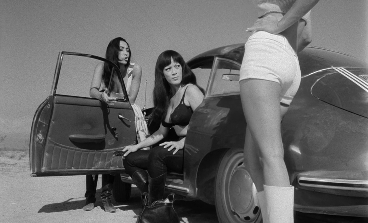 What Is Exploitation Cinema? Definition & Key Examples 8 Scene from Faster, Pussycat! Kill! Kill! showing three women by a car in the desert, dressed in revealing clothes and striking bold poses.