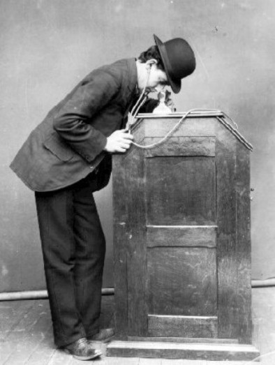 What Is a Silent Movie? Definition & History Explained 2 Man in a bowler hat leaning over a wooden Kinetoscope, watching a short film through a peephole