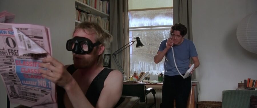 What is a Romcom? Definition, History & Tropes 3 Hugh Grant on the phone while his roommate, in goggles and a vest, reads a newspaper.