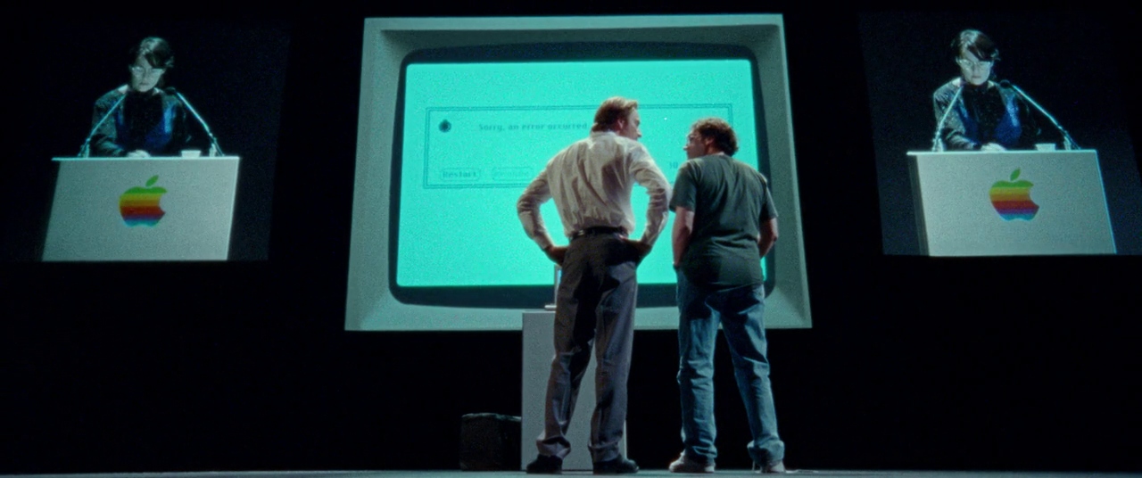 What Is a Biopic? Definition & Film Examples 4 Steve Jobs stands on stage with a giant computer screen behind him while two men argue in front of it during a product launch.
