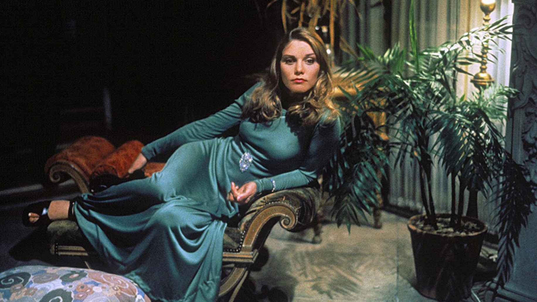 What Are Sexploitation Films? Definition, Movies & Global Impact 12 Woman in a long green dress lounging on a velvet bench beside a potted plant, looking off-camera