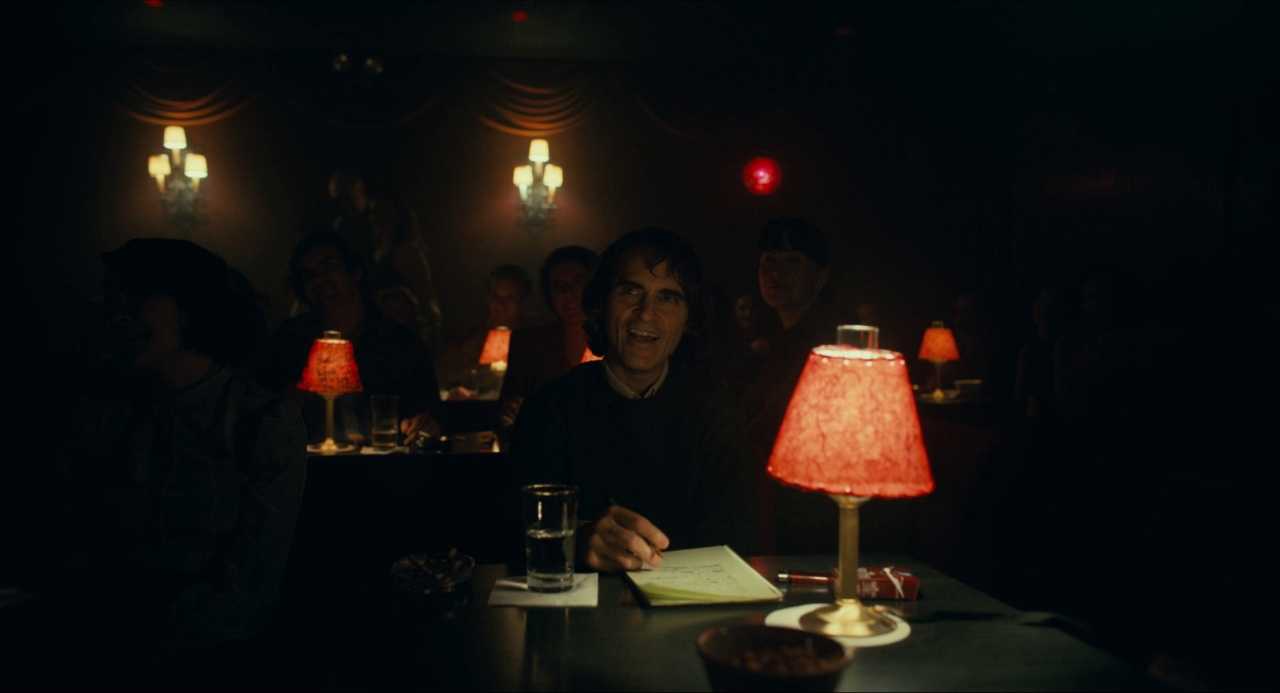 What Does a Cinematographer Do? Job Description & Real DP Examples 5 Arthur Fleck sits in a dim nightclub lit by red table lamps, his face softly lit in a warm amber glow.