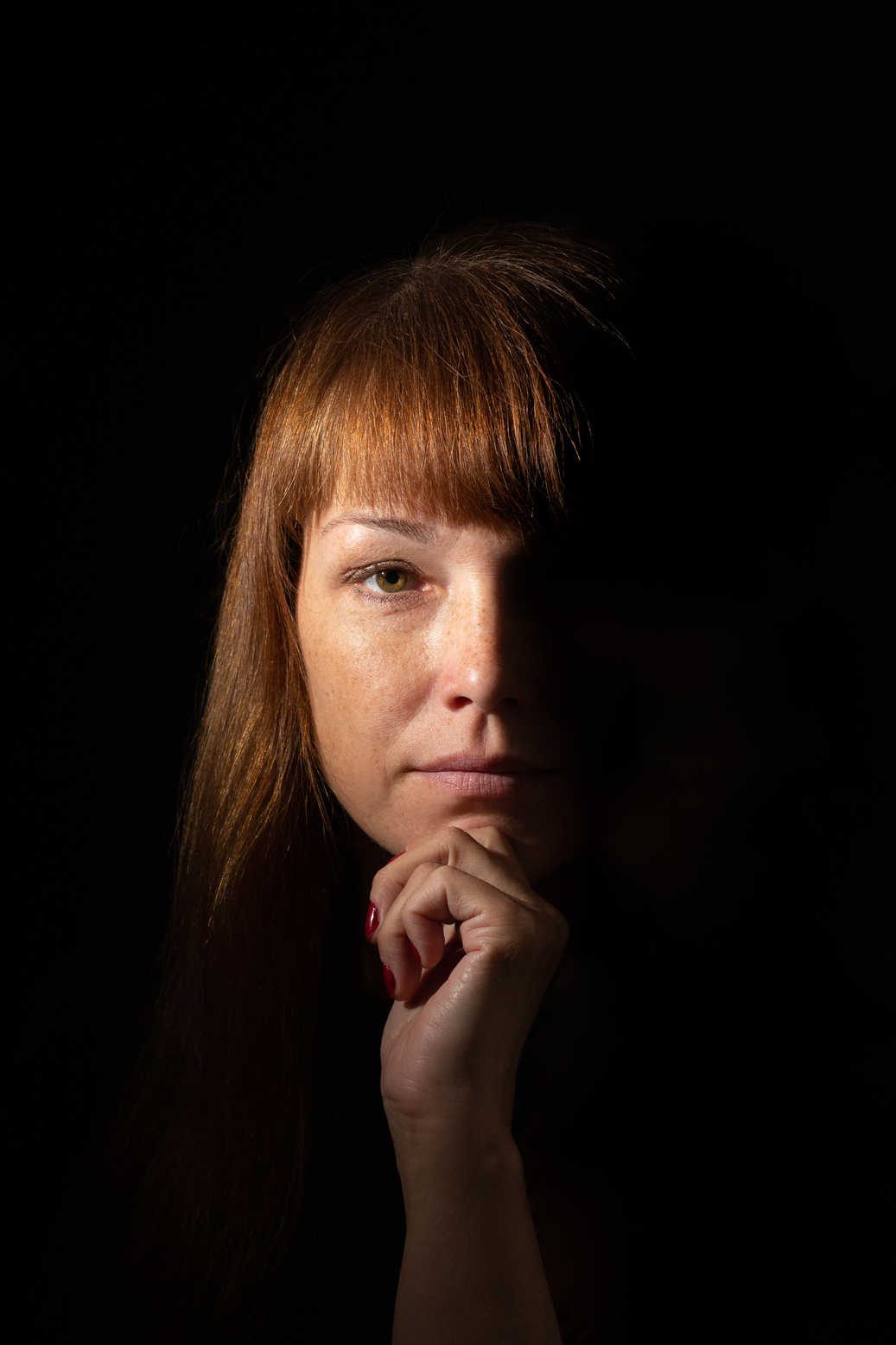 Portrait Lighting Setup Guide: Rembrandt, Loop & Beyond 3 Portrait of a woman lit with 1-point side light (split lighting), showing one half of her face and hand illuminated while the other half remains in shadow against a dark background.