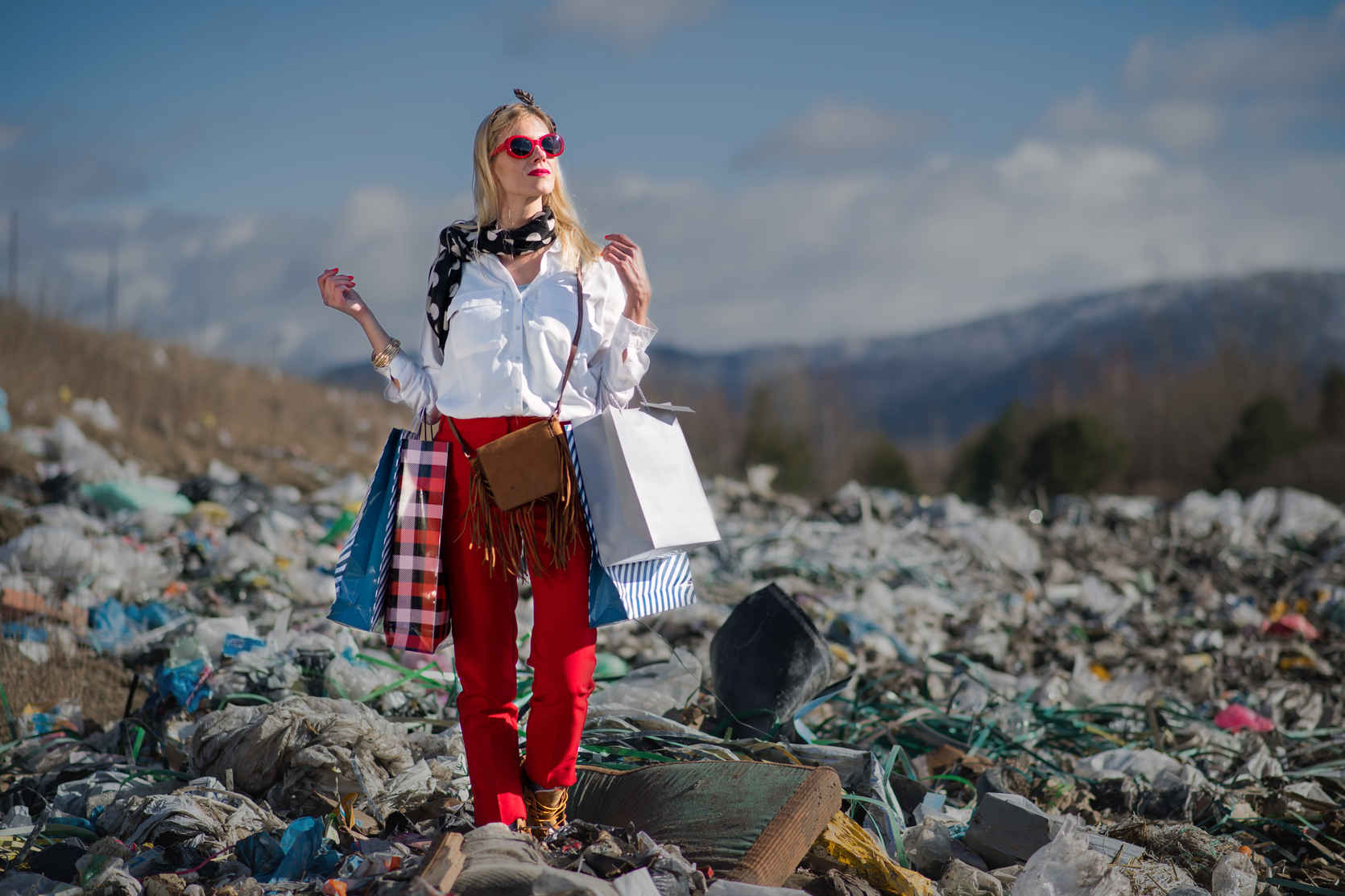 What Is Conceptual Photography? Definition, Style & Examples 4 A fashionable woman stands confidently with shopping bags in a landfill, wearing bright red pants and sunglasses against a backdrop of trash and mountains.