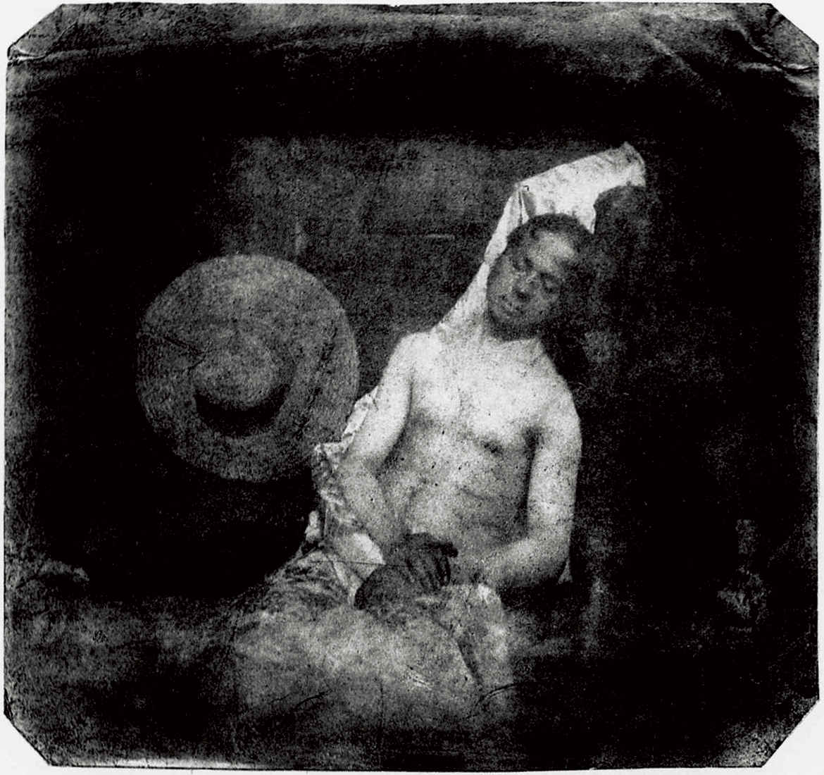 What Is Conceptual Photography? Definition, Style & Examples 7 Staged black-and-white photograph of a shirtless man posed as if dead, with closed eyes and hands folded, leaning against a wall beside a wide-brimmed hat.