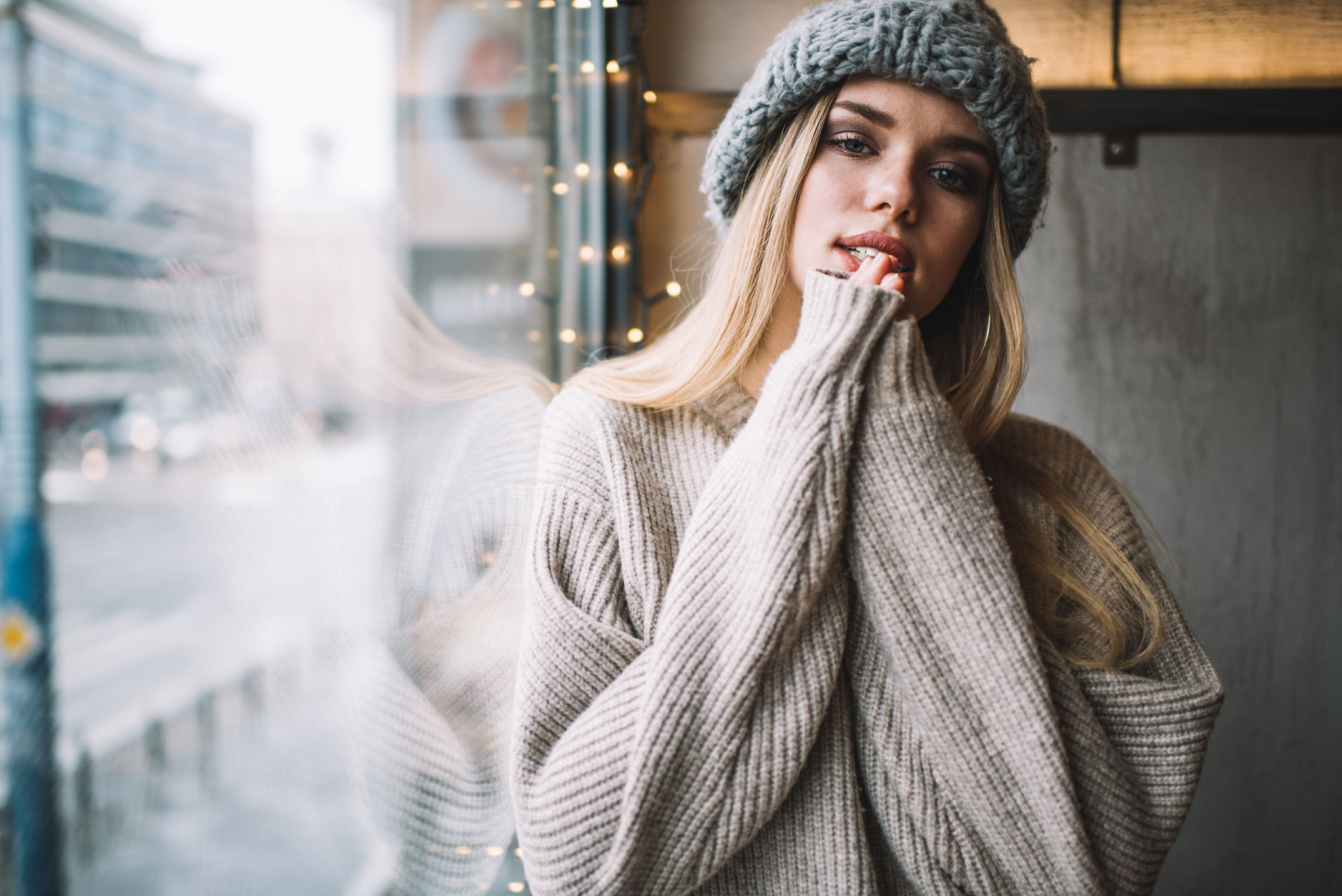 What Is Brand Photography? Definition, Uses & Planning Steps 3 Young woman in knit sweater and beanie poses by a window with fairy lights, creating a cozy winter lifestyle scene