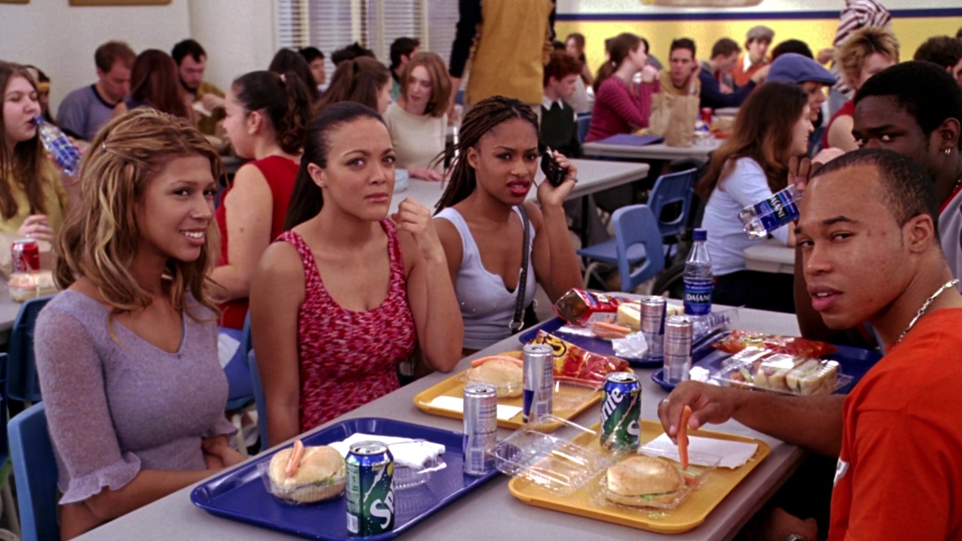 What Is an Overstatement? Definition & Examples from Film 4 A group of high school students sit at a cafeteria table with lunch trays, sandwiches, and soda cans, looking toward the camera while other students fill tables in the background.