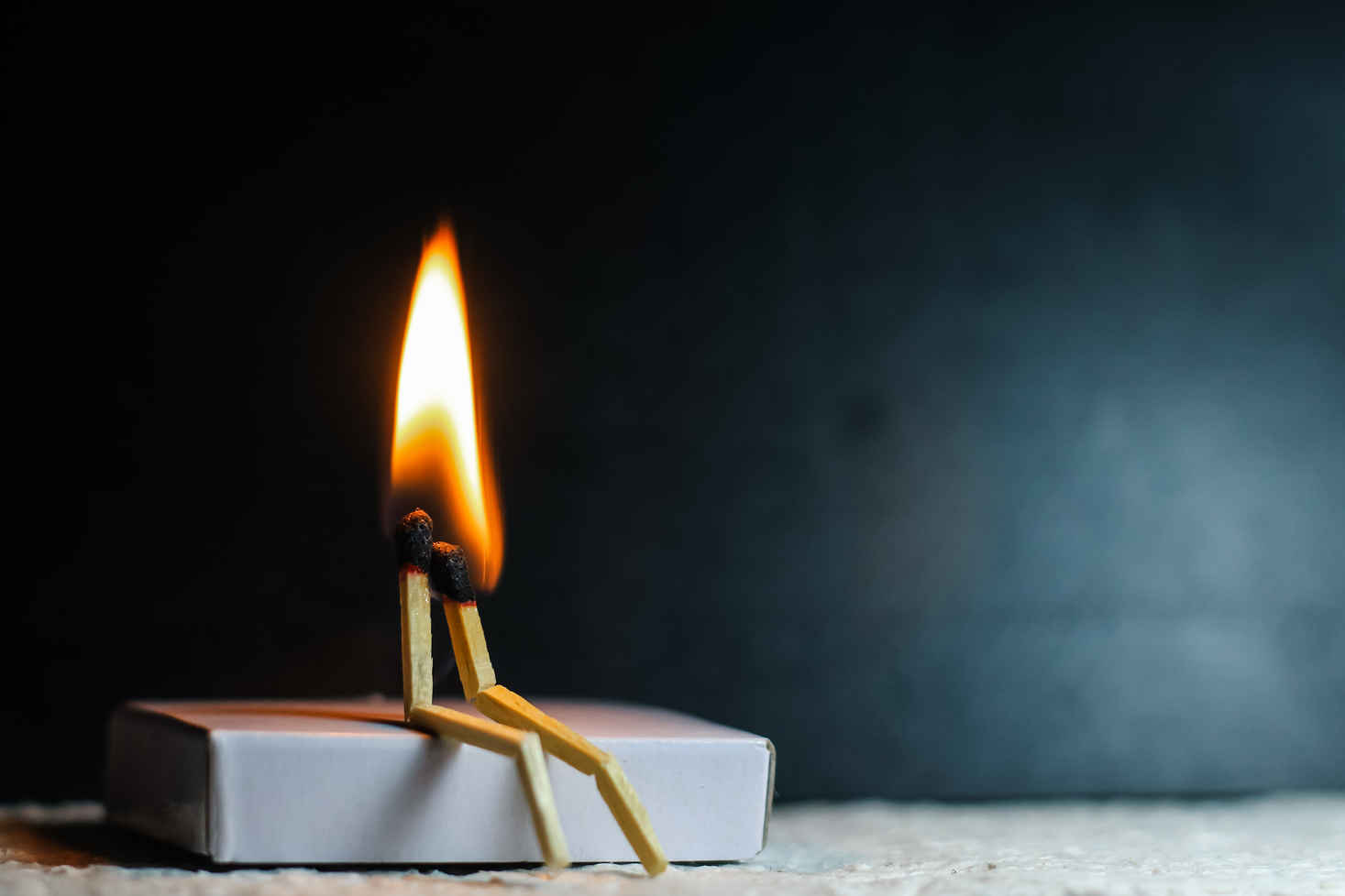 What Is Conceptual Photography? Definition, Style & Examples 6 Two burnt matchsticks propped up like a couple sitting together, joined at the flame, with a dark background and soft lighting.