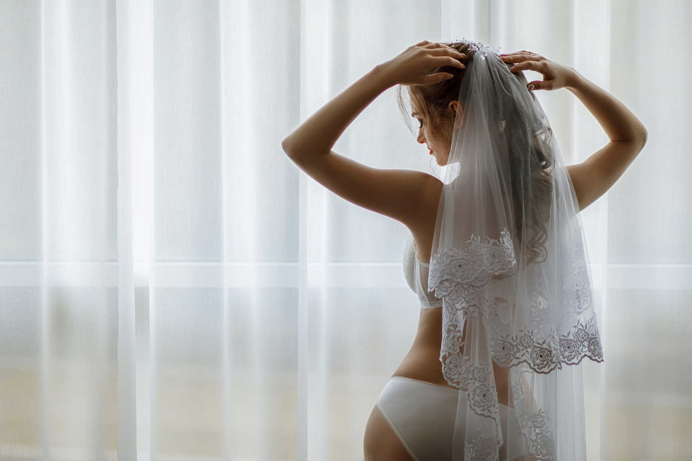 What Is Glamour Photography? Definition & Complete Guide to Style 9 Bride in lingerie and veil adjusts her hair in front of a softly lit window