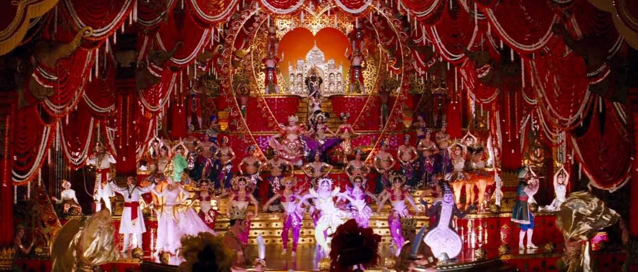 What Is an Overstatement? Definition & Examples from Film 2 Wide shot of the Moulin Rouge stage packed with dancers in colorful costumes, surrounded by red curtains, gold set pieces, and bright stage lights.